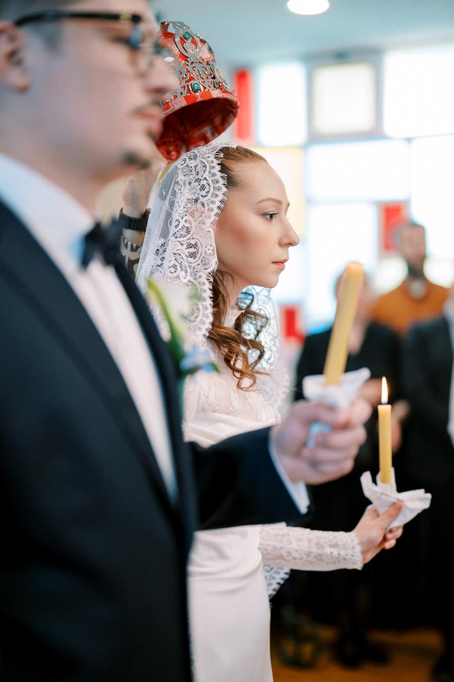 A bride in a lace veil holds a candle during a traditional wedding ceremony, with an ornate crown being placed on her head. A groom stands beside her, also holding a candle, in a brightly lit church.