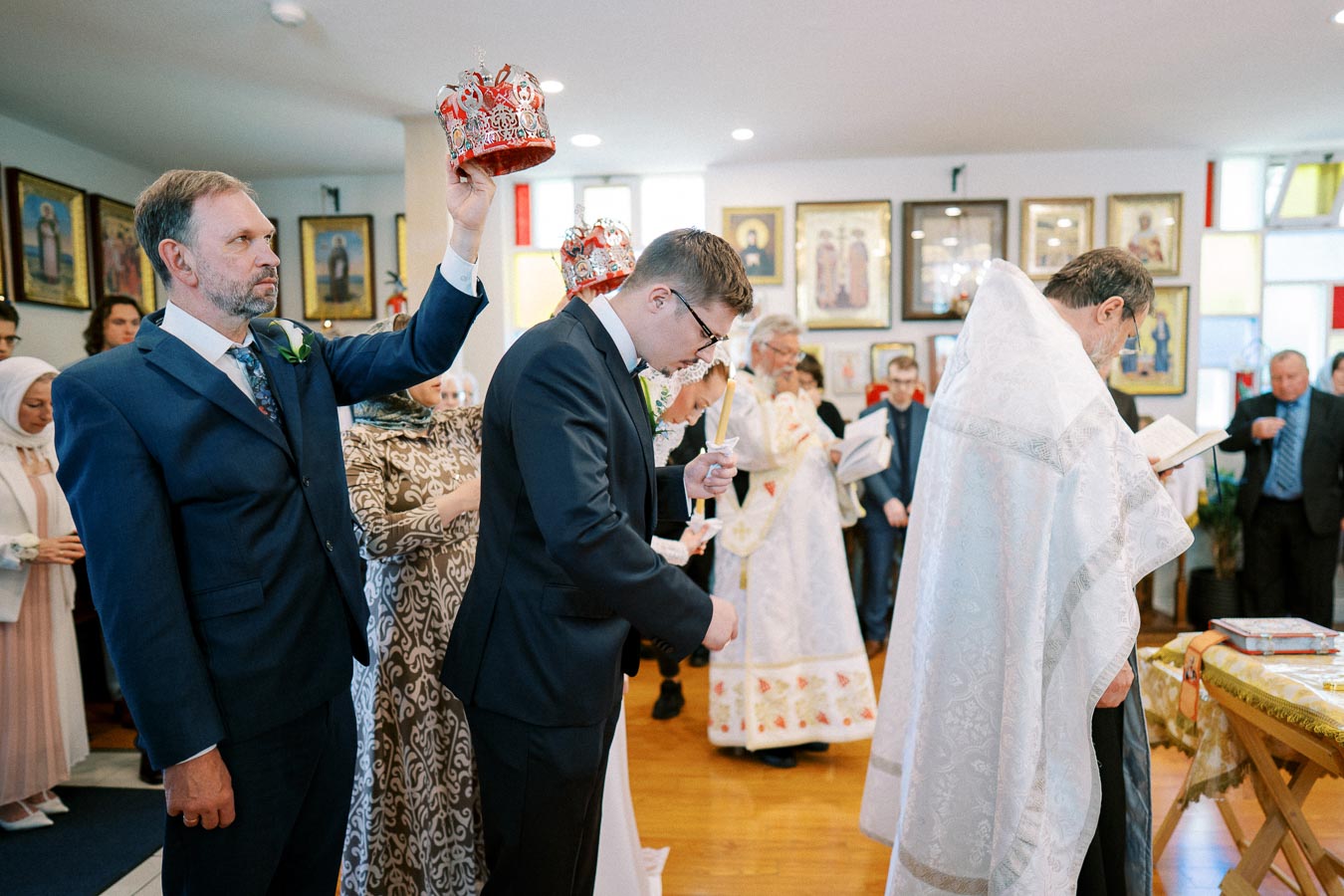 Orthodox wedding ceremony with guests in traditional attire, featuring a priest in white robes and a groom holding a candle, centered in a church with religious iconography and crowns being placed by an attendant.