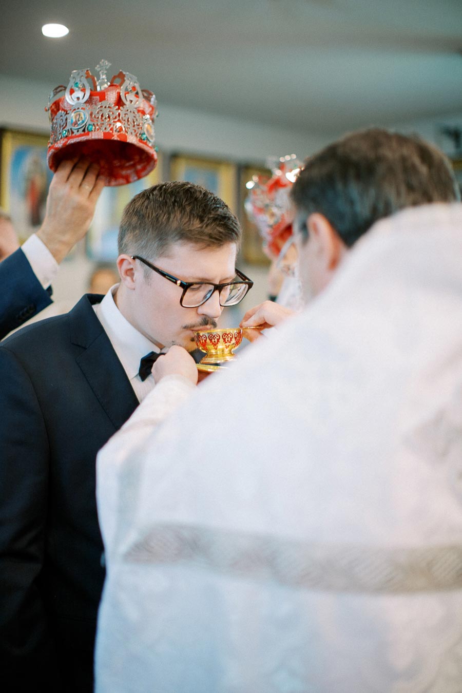 A groom in a formal suit receiving a ceremonial blessing during an Eastern Orthodox wedding, with a priest holding a chalice and a crown being placed above his head.