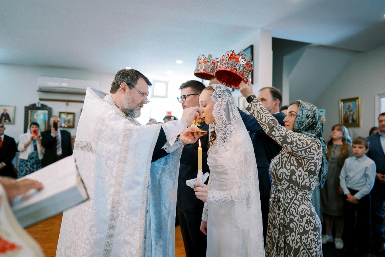 Orthodox wedding ceremony with bride and groom crowned, a priest serving communion, and guests observing in a church setting.
