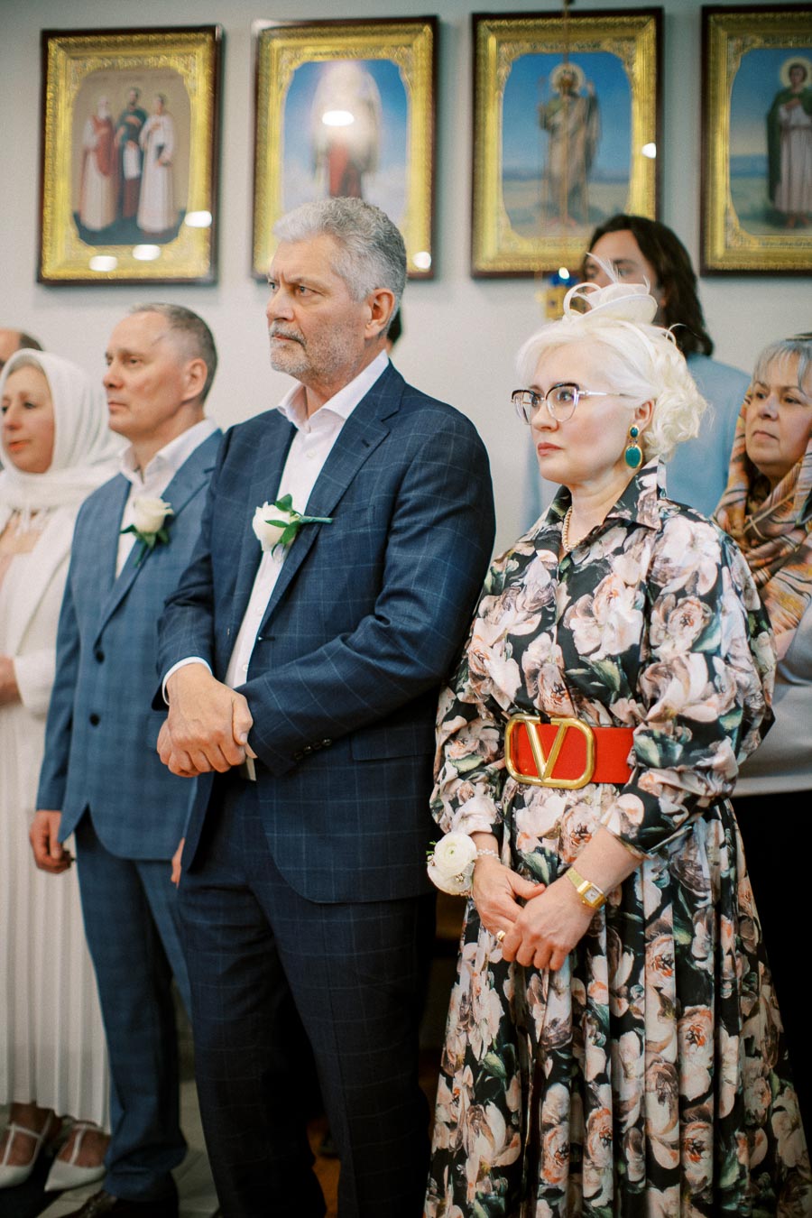 A group of people standing indoors, dressed formally with floral and checkered patterns, holding white flowers, against a backdrop of religious paintings.
