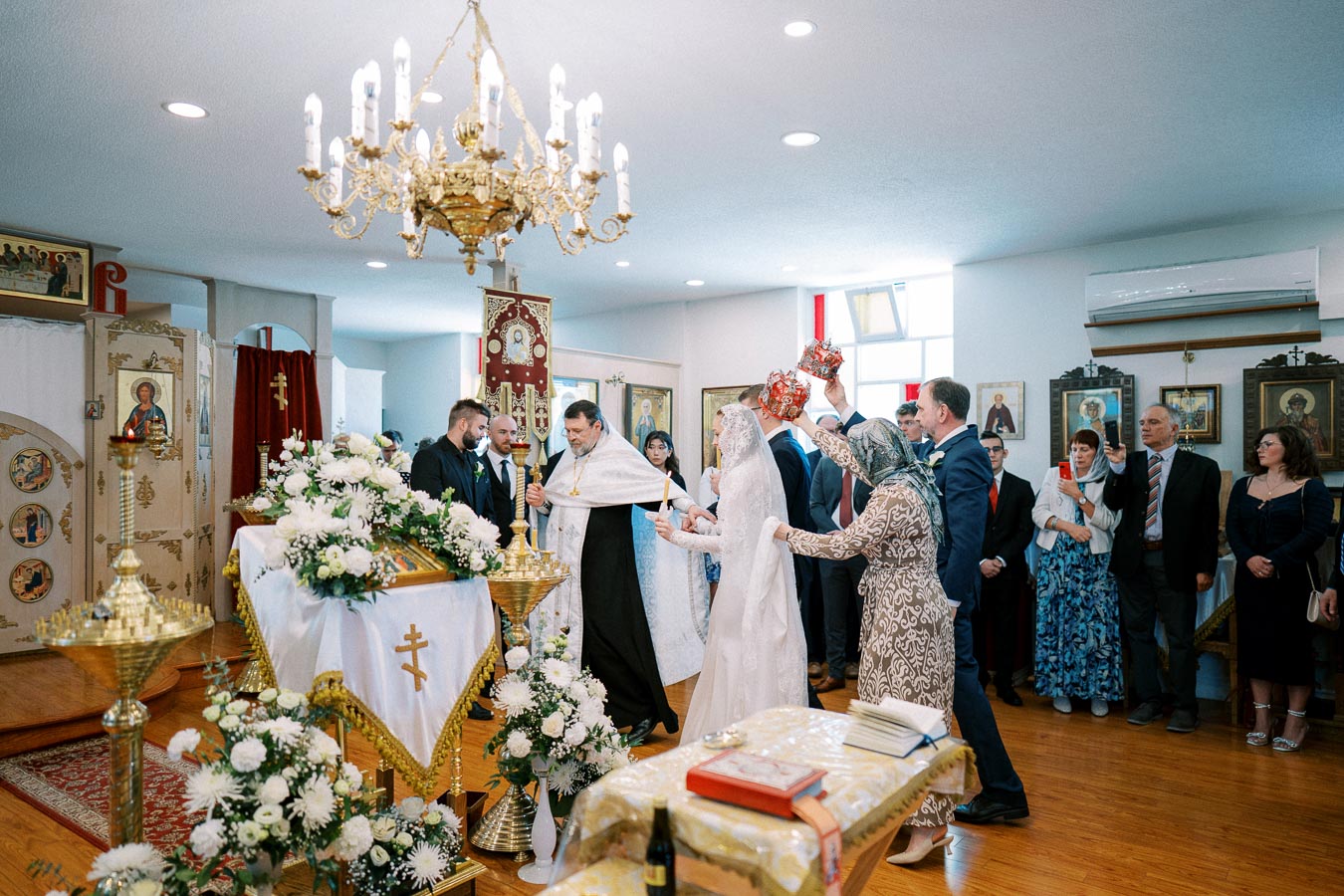 Orthodox wedding ceremony inside a beautifully decorated church, featuring bride and groom participating in traditional rituals with a priest, surrounded by family and friends, under an ornate chandelier.