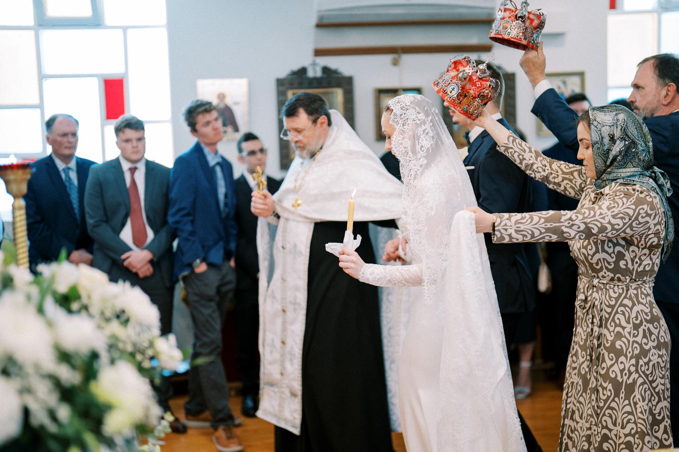 Orthodox wedding ceremony in a church, featuring a bride in a white lace gown holding a candle, surrounded by guests and clergy, with ornate crowns being held over the couple's heads.
