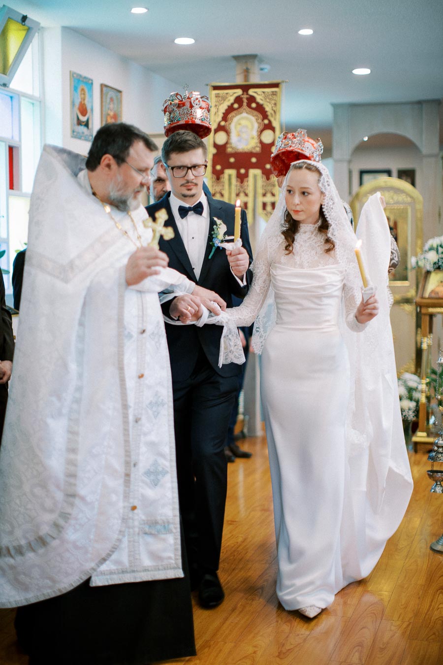 Orthodox wedding ceremony with a bride and groom wearing crowns, holding candles, led by a priest in traditional vestments inside a church.