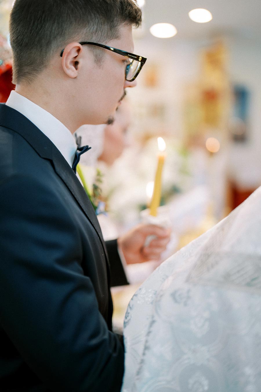 Groom in a suit holding a candle during a wedding ceremony, captured in a softly lit indoor setting.