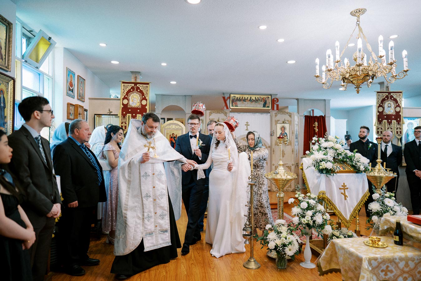 Orthodox wedding ceremony with a bride and groom holding candles, surrounded by clergy and guests in a church decorated with religious icons and floral arrangements.