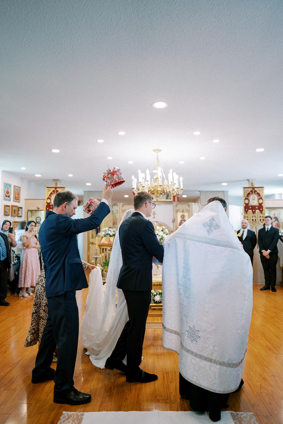 A traditional religious wedding ceremony inside a church, featuring a couple standing together at the altar. A priest or officiant is present, and a man in a suit holds ornate crowns over the couple's heads, symbolizing an important ritual. The church interior is adorned with icons and decorations, adding to the solemn atmosphere.