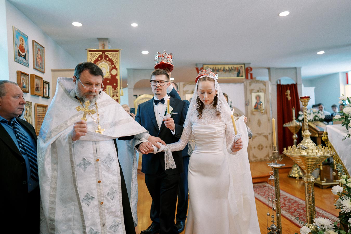 Orthodox wedding ceremony with bride and groom holding candles, led by a priest in traditional robes, surrounded by religious icons and ceremonial decor.