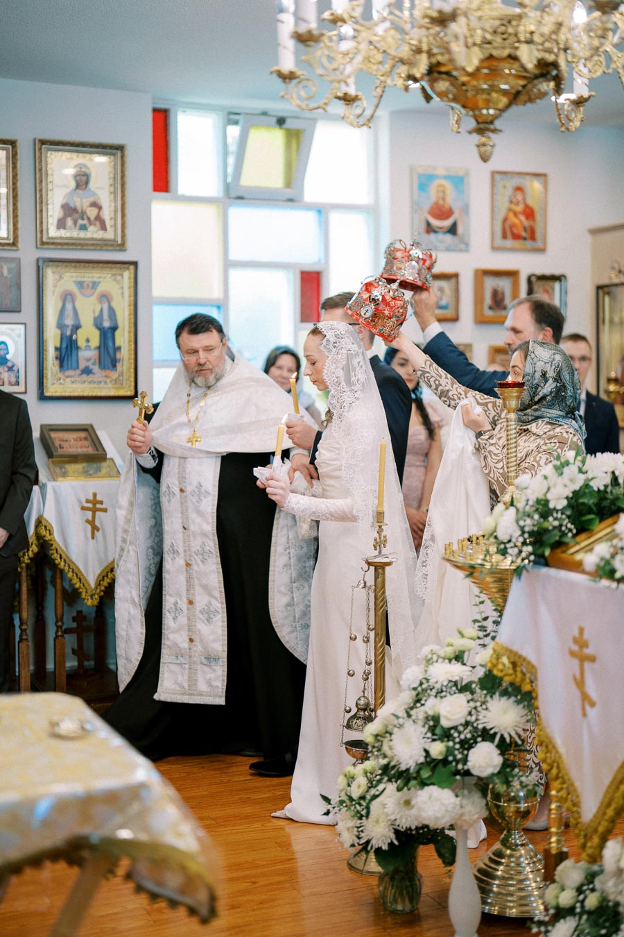 Orthodox wedding ceremony with priest and couple, adorned with crowns, in a beautifully decorated church setting.