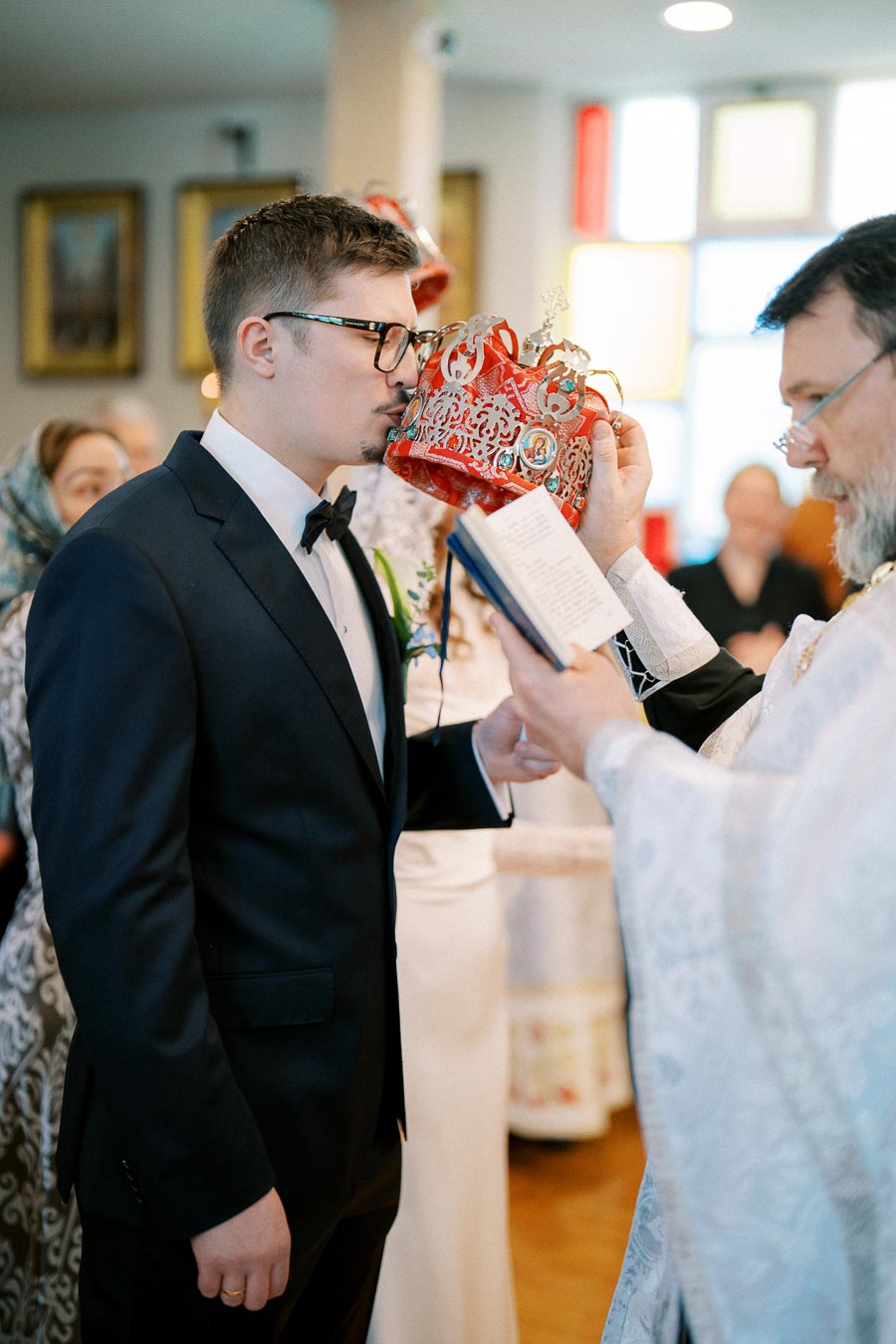 A groom in a formal suit and glasses participates in a religious wedding ceremony, kissing a red ornamental crown held by a priest in ceremonial attire, with a congregation in the background.