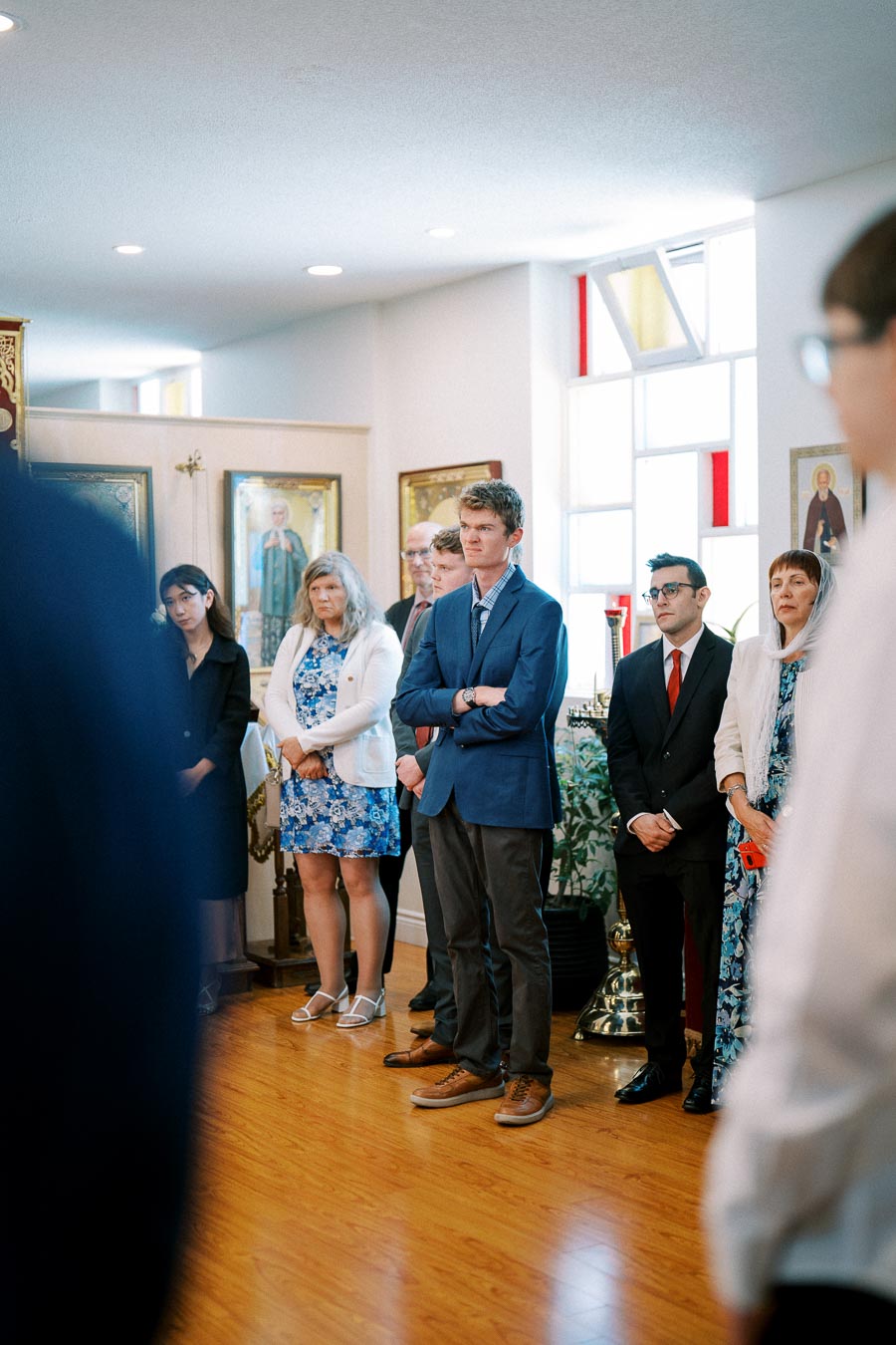 A group of people attentively standing inside a brightly lit room with wooden floors, featuring religious icons on the walls and large windows allowing natural light.