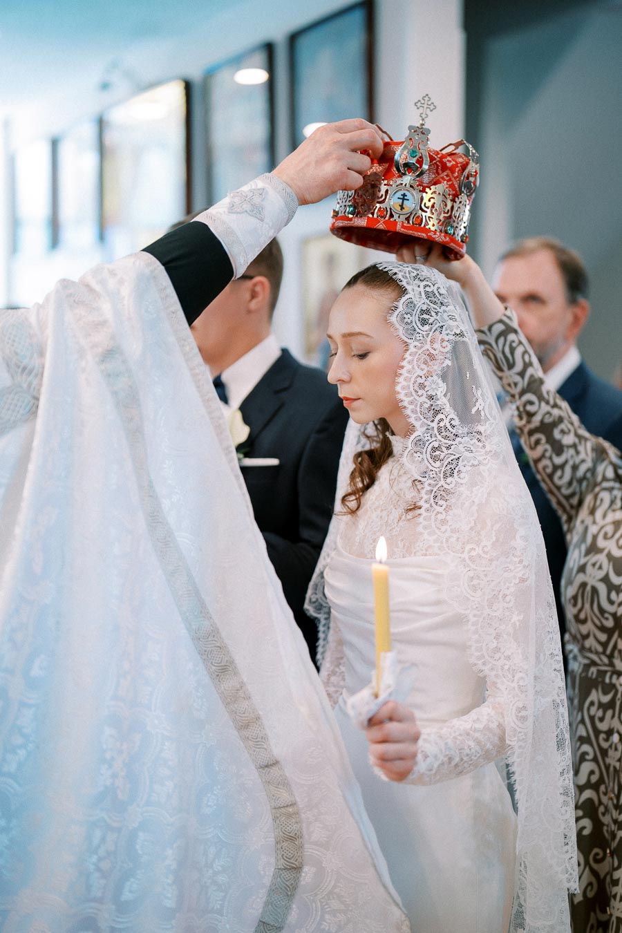 A bride in a traditional lace wedding dress and veil holds a candle during a ceremony. A priest in white vestments places an ornate crown on her head, symbolizing a religious marriage ritual.
