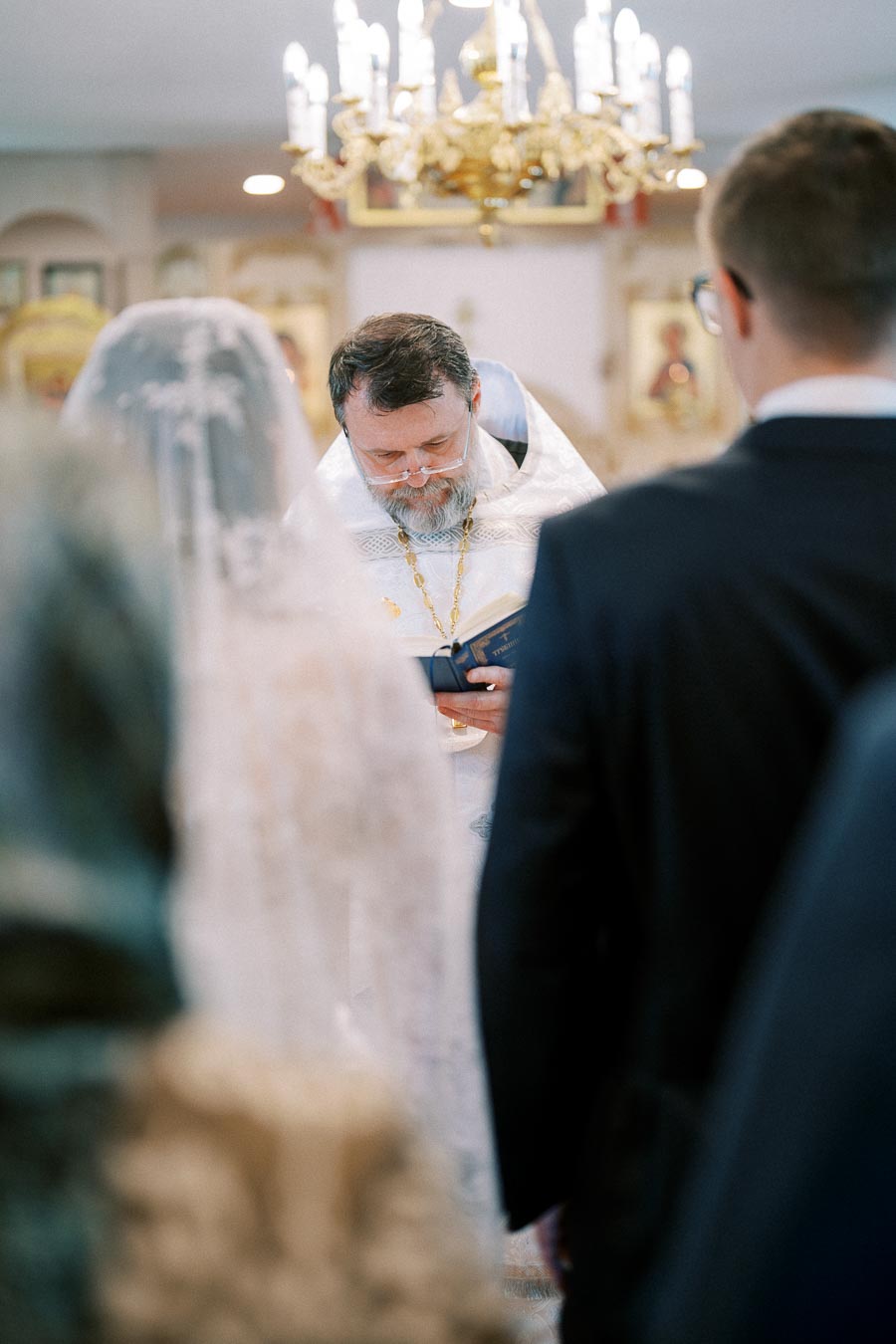 An officiant in white robes leading a wedding ceremony in a church, with the bride in a veil and the groom in a suit in the foreground, under an ornate chandelier.