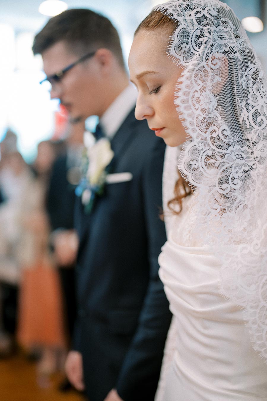 A bride in a lace veil and groom in a black suit standing close together, captured in a serene wedding ceremony atmosphere.