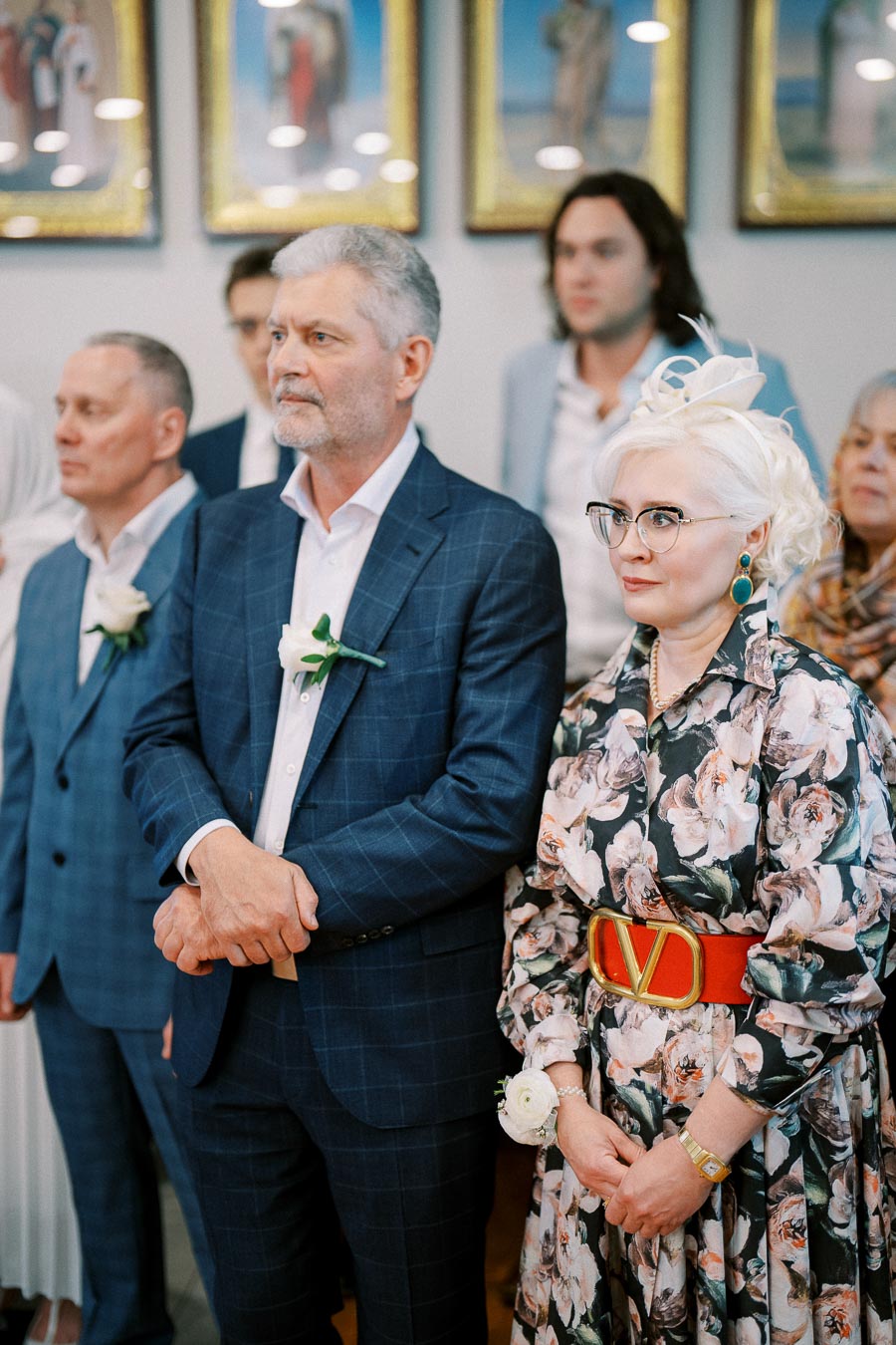 A group of elegantly dressed people attending a formal event, with the focus on a man in a blue checkered suit and a woman in a floral dress with a red belt, standing in a well-lit room with framed art on the walls.