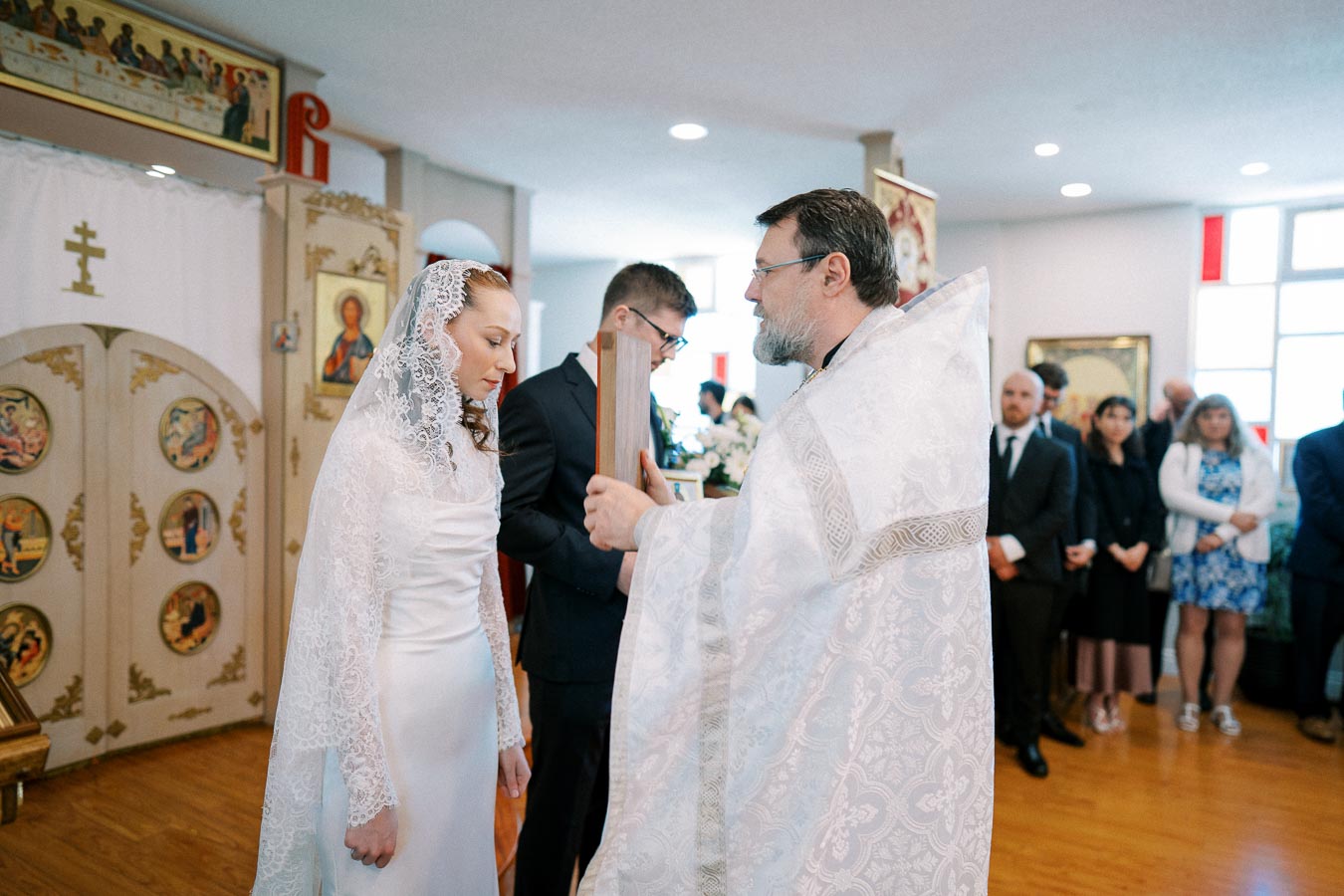 Orthodox wedding ceremony with bride in lace veil and priest conducting ritual in ornately decorated church, surrounded by guests.