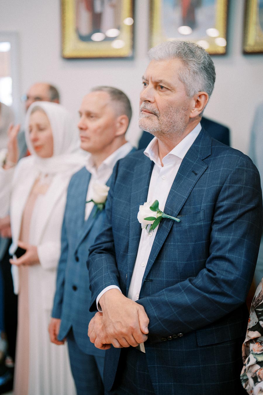 A group of formally dressed individuals attending a ceremony, with two men in blue suits wearing white boutonnieres and a woman in a white headscarf, standing attentively in a decorated room.