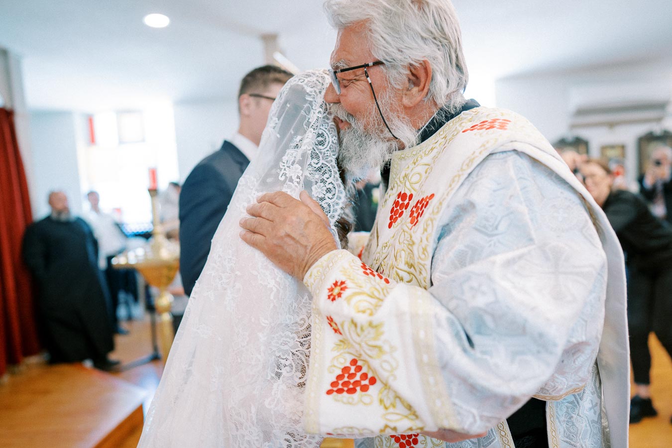 Elderly priest embracing a bride in a lace veil during a traditional wedding ceremony in a church setting.