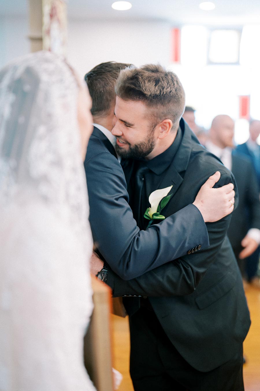 Wedding ceremony moment with groom hugging a smiling guest, bride partially visible in the foreground, capturing joyful emotions and elegant attire indoors.