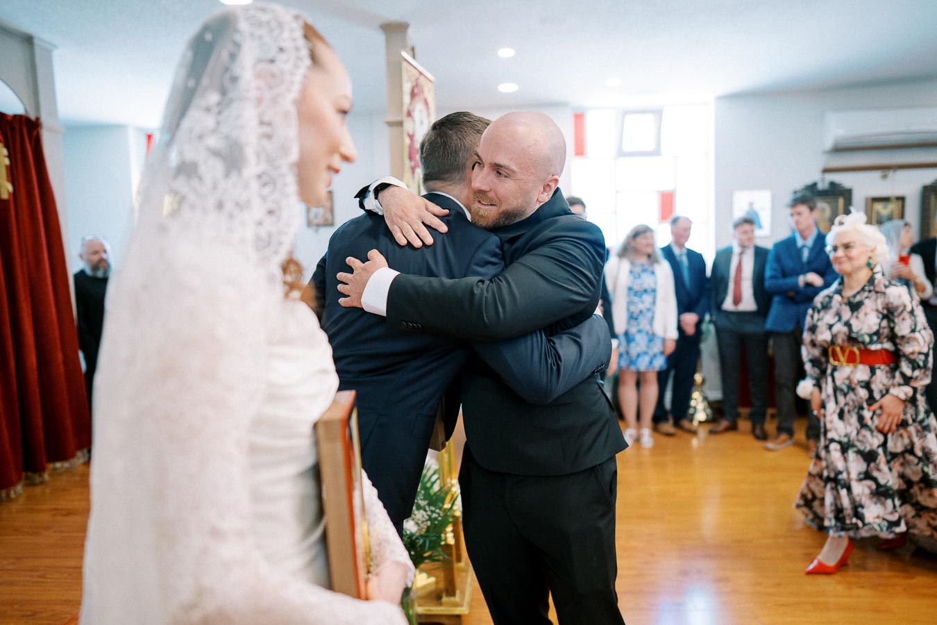 A groom hugging a guest during a wedding ceremony in a church, with a bride holding a religious icon in the foreground, surrounded by attendees dressed in formal wear.