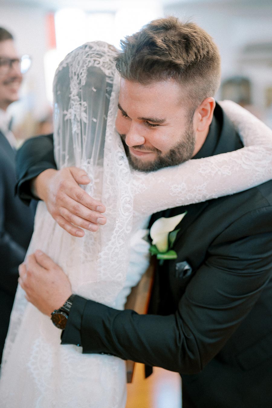 Emotional wedding scene depicting a groom in a black suit warmly embracing the bride who's wearing a lace veil, capturing a heartfelt moment of joy.