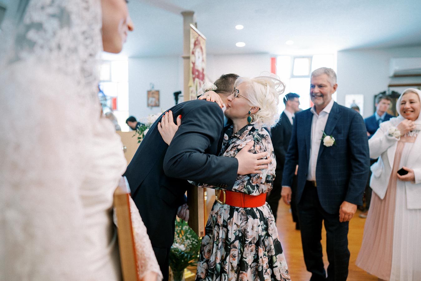 A joyful wedding reception scene with a woman embracing a man in a suit, surrounded by smiling guests in a warmly lit venue, highlighting celebration and togetherness.