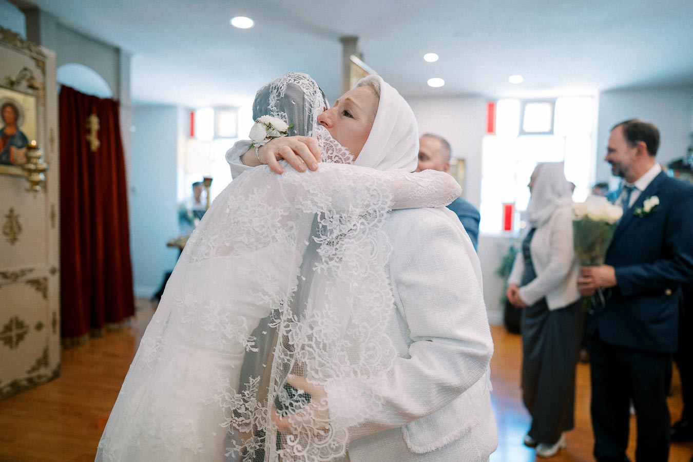A bride in a lace wedding dress embraces a woman in a white headscarf at a traditional ceremony, surrounded by family and friends, in a warmly lit room with religious icons.
