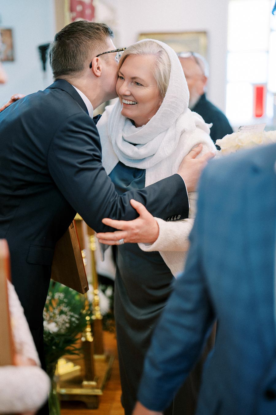 A joyful woman in a white headscarf smiles while embracing a man in a suit during a celebration or ceremony, with elegant decor in the background.