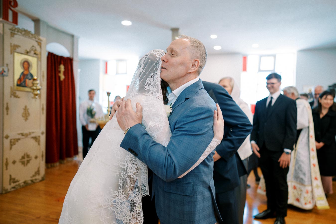 A bride in a white lace dress hugs an older man in a blue suit during a church wedding ceremony, surrounded by guests and a priest in the background.