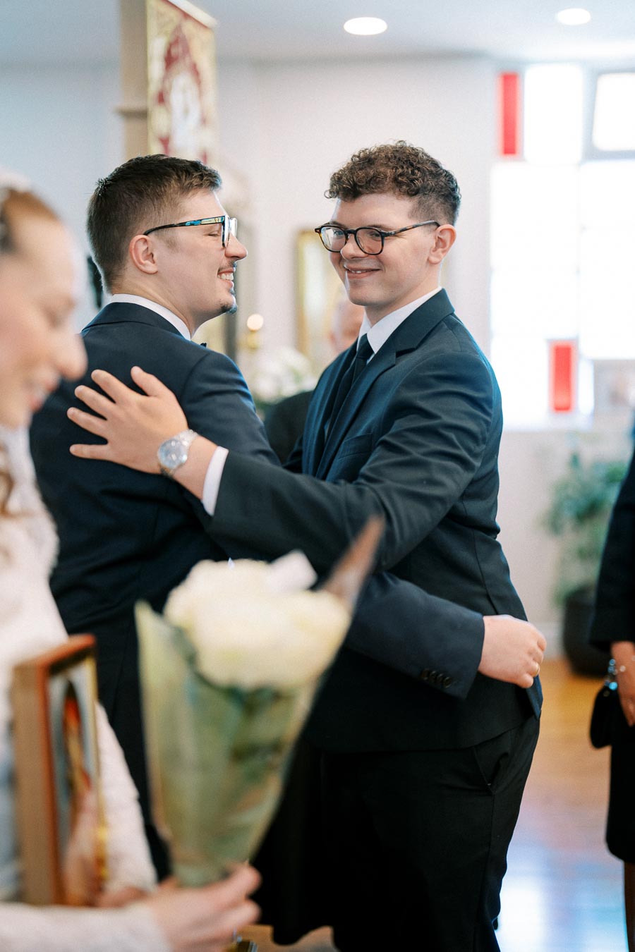 Two men in formal suits smiling and embracing at a wedding ceremony, surrounded by a joyful atmosphere with a person holding a bouquet in the foreground.
