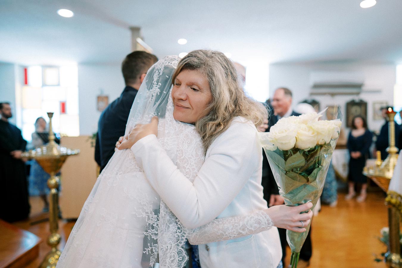 A bride in a lace wedding gown receives a heartfelt hug from a woman holding a bouquet of white roses at an indoor wedding ceremony.