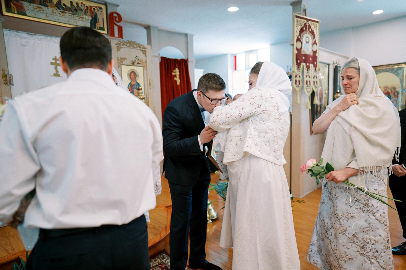 A traditional Orthodox church ceremony with a man in a suit kissing a child's forehead held by a woman in white lace, surrounded by religious icons and attendees wearing headscarves, holding flowers.