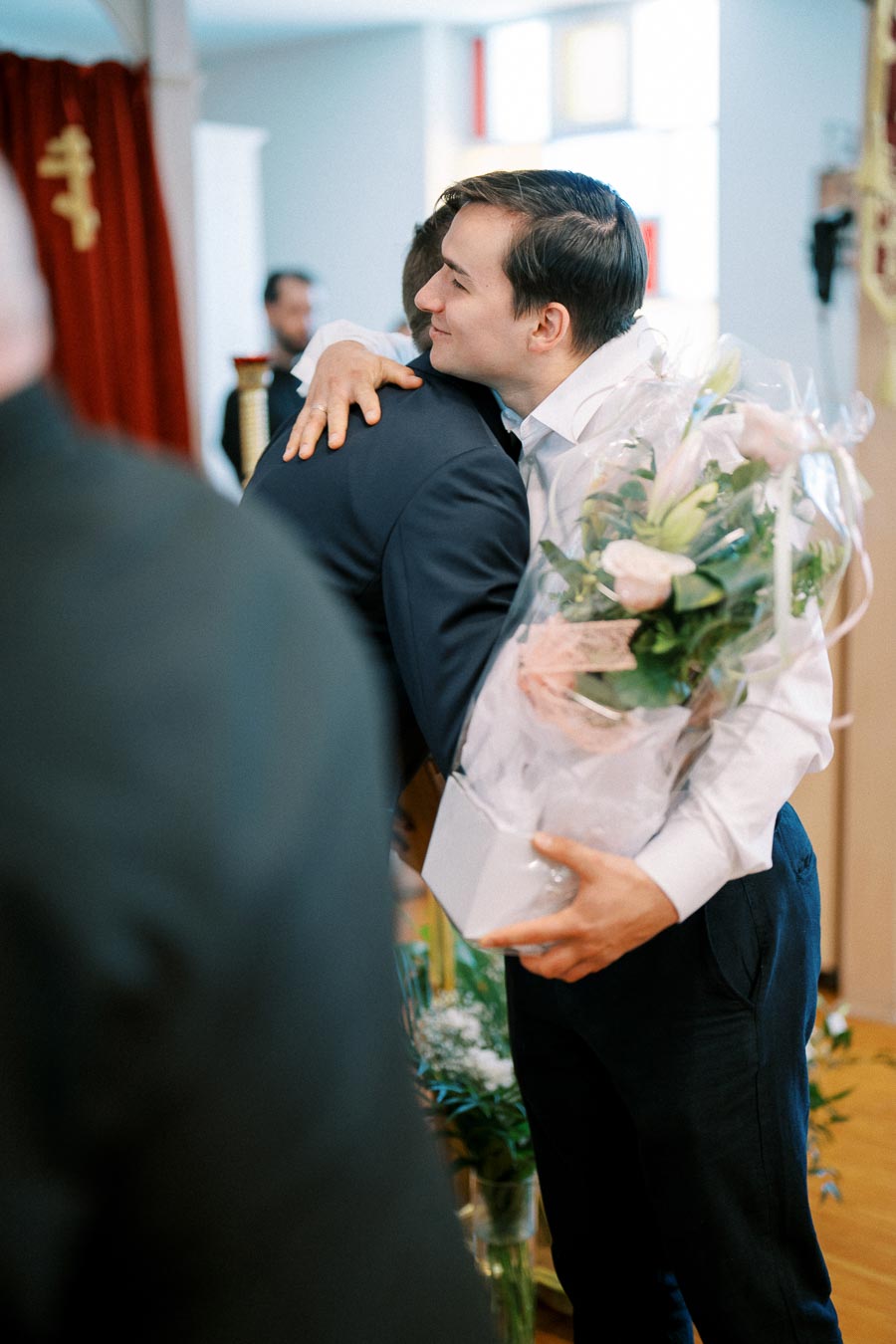 Two men embracing during a joyful celebration, one holding a bouquet of flowers wrapped in plastic, suggesting a warm and festive occasion indoors.