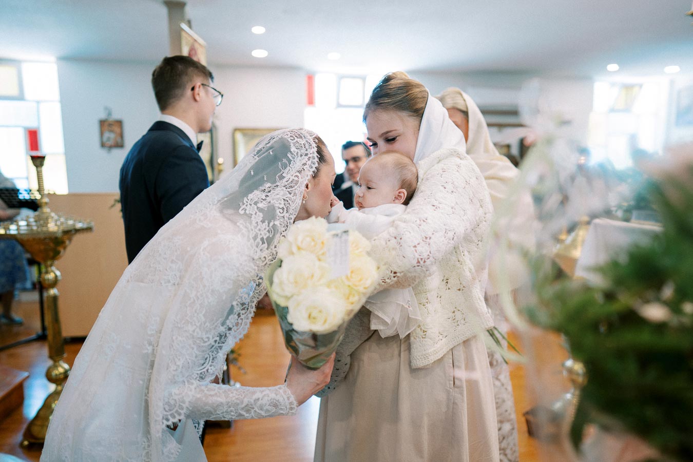 A christening ceremony inside a church, where a woman in a white lace veil leans forward to kiss a baby held by another woman in a white headscarf. The setting is adorned with flowers and religious decor.