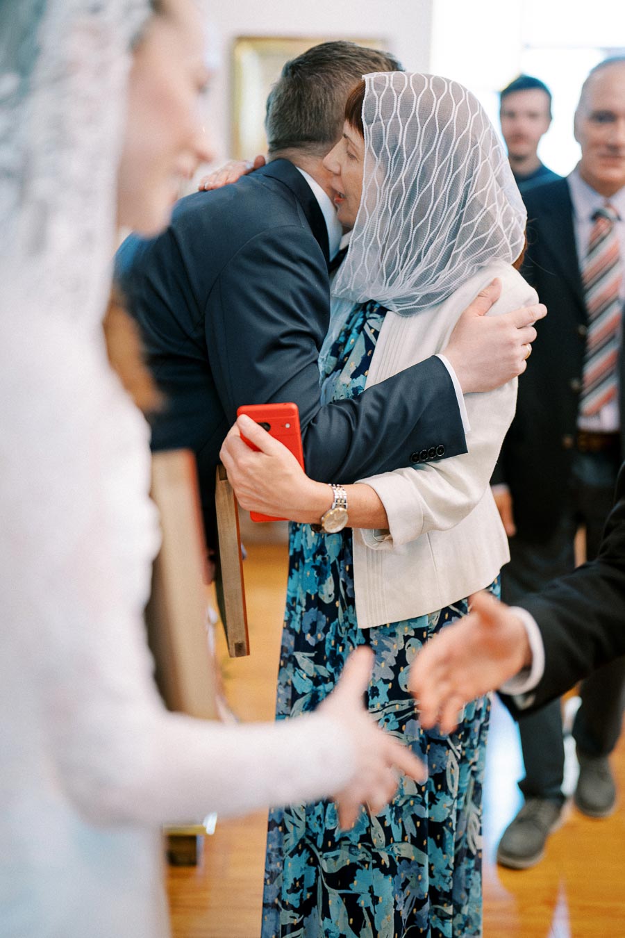 A woman wearing a veil and blue floral dress hugs a man in a suit, holding a red phone. Others stand in the background, and a hand extends in the foreground, suggesting a warm gathering, possibly at a wedding or celebration.