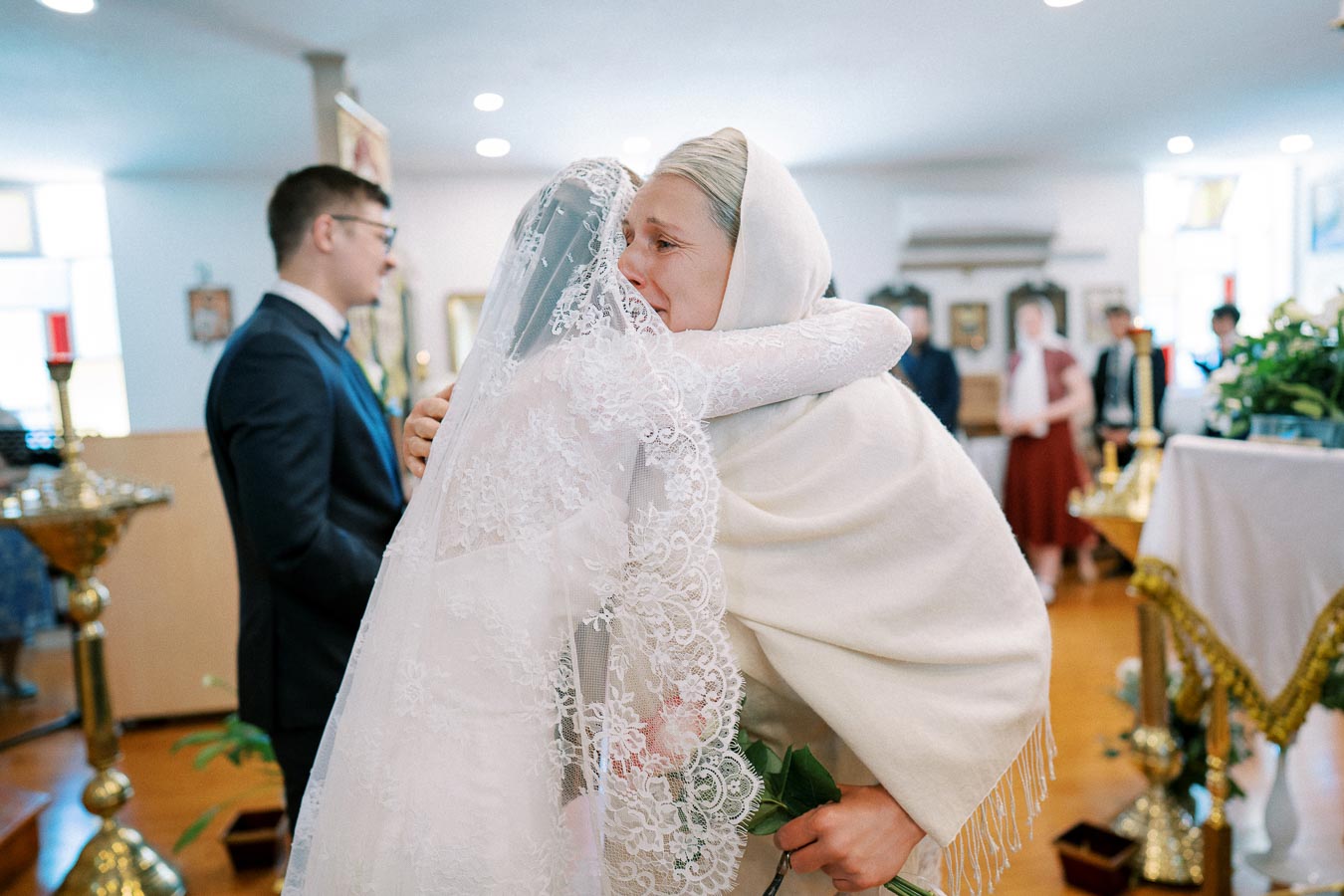 A bride in a lace veil embraces an older woman in white during an emotional wedding ceremony, with a groom and guests in soft focus in the background, surrounded by traditional decor.