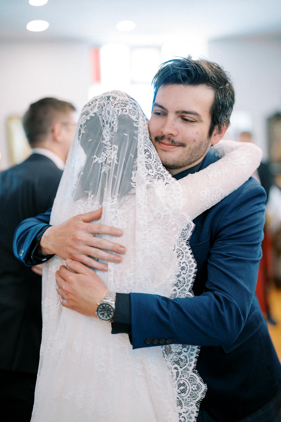 A groom in a blue suit warmly embraces a bride wearing a lace veil during their wedding ceremony, conveying love and joy.