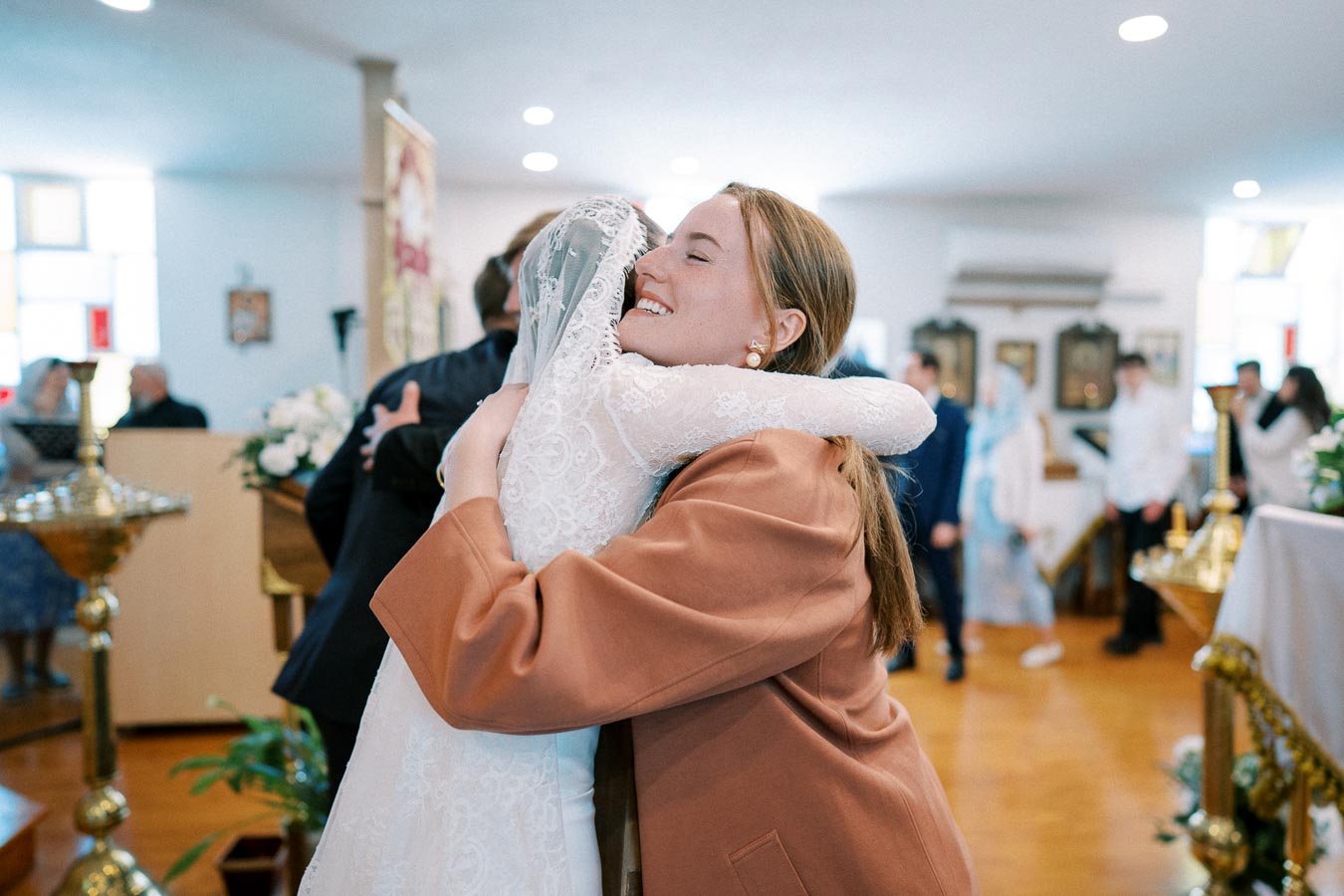 A bride in an elegant lace wedding dress embraces a friend joyfully inside a beautifully decorated church during a wedding ceremony.