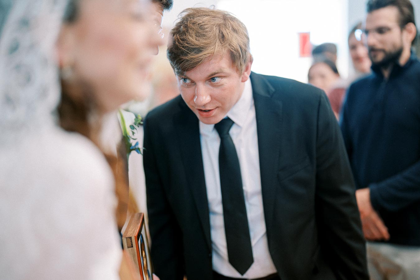Man in a suit leaning forward and speaking at an indoor event, with blurred individuals in the background.