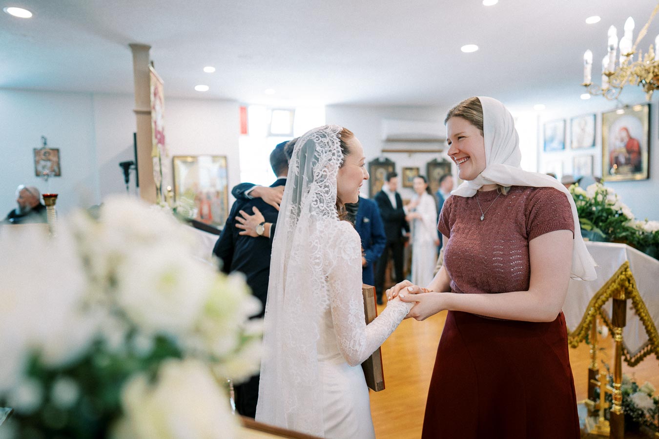 Two women smiling and holding hands in a warmly-lit interior, possibly a wedding or celebration, with a focus on flowers in the foreground and framed pictures on the walls.