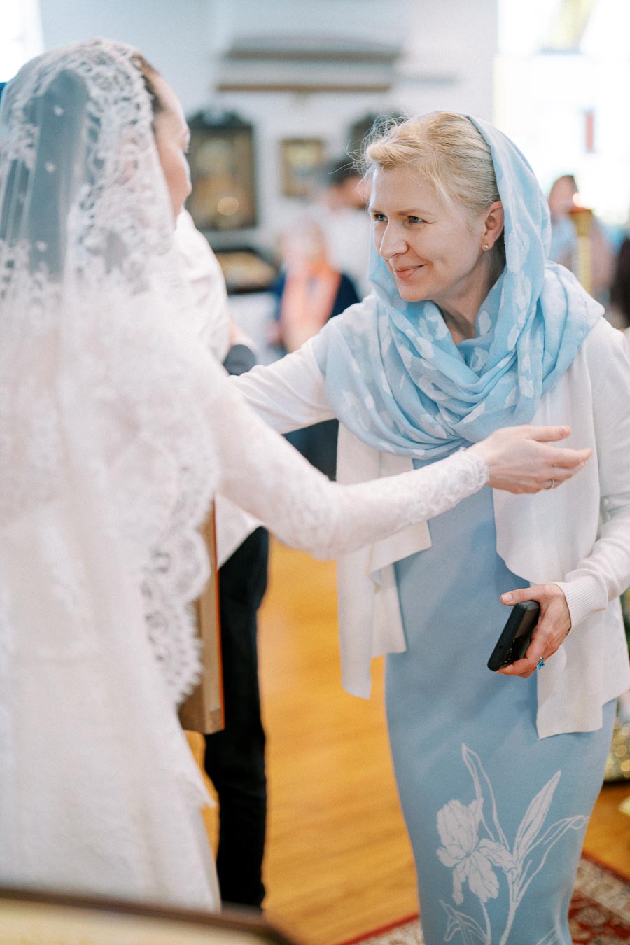 A woman in a blue headscarf smiles warmly while greeting a bride in a lace wedding dress indoors.