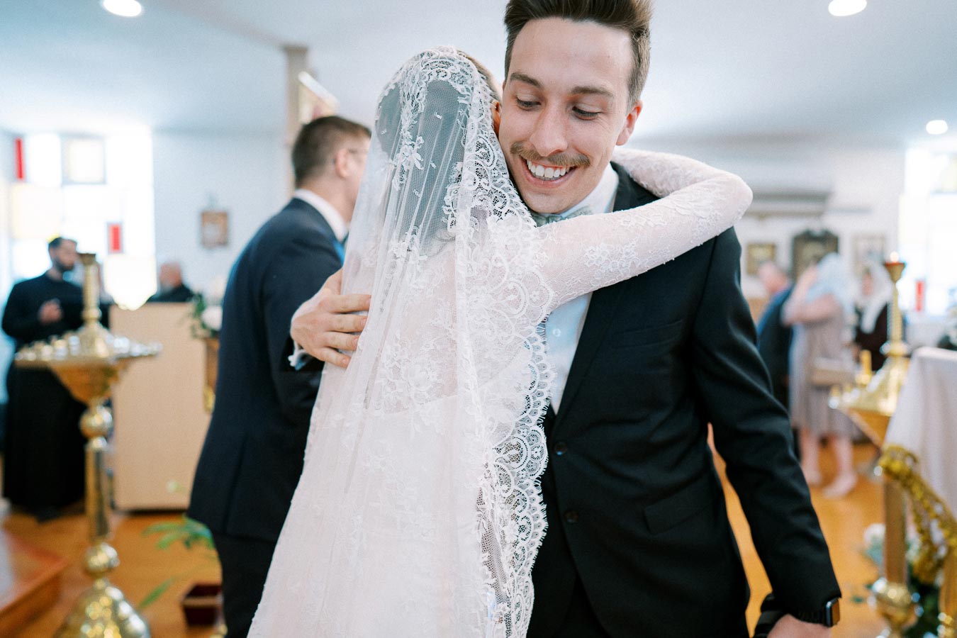 A groom smiling as he hugs his bride in a lace veil during a wedding ceremony in a beautifully decorated church setting.