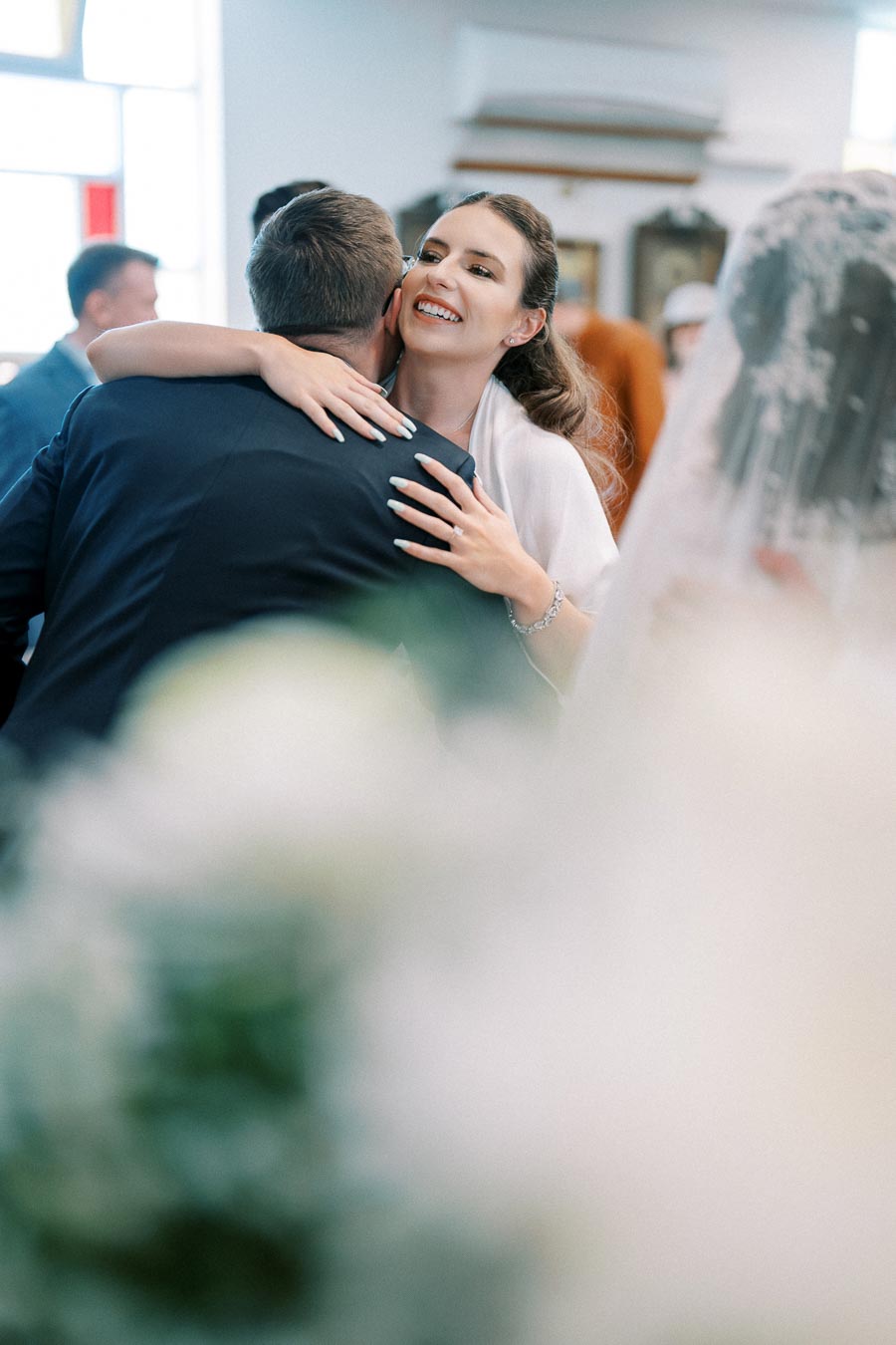 A bride warmly embraces a guest, smiling joyfully, inside a beautifully decorated venue during a wedding celebration.