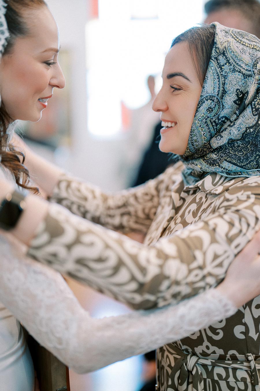 Two women embracing warmly, with one wearing a patterned headscarf and the other in a white lace outfit, engaged in a joyful conversation.