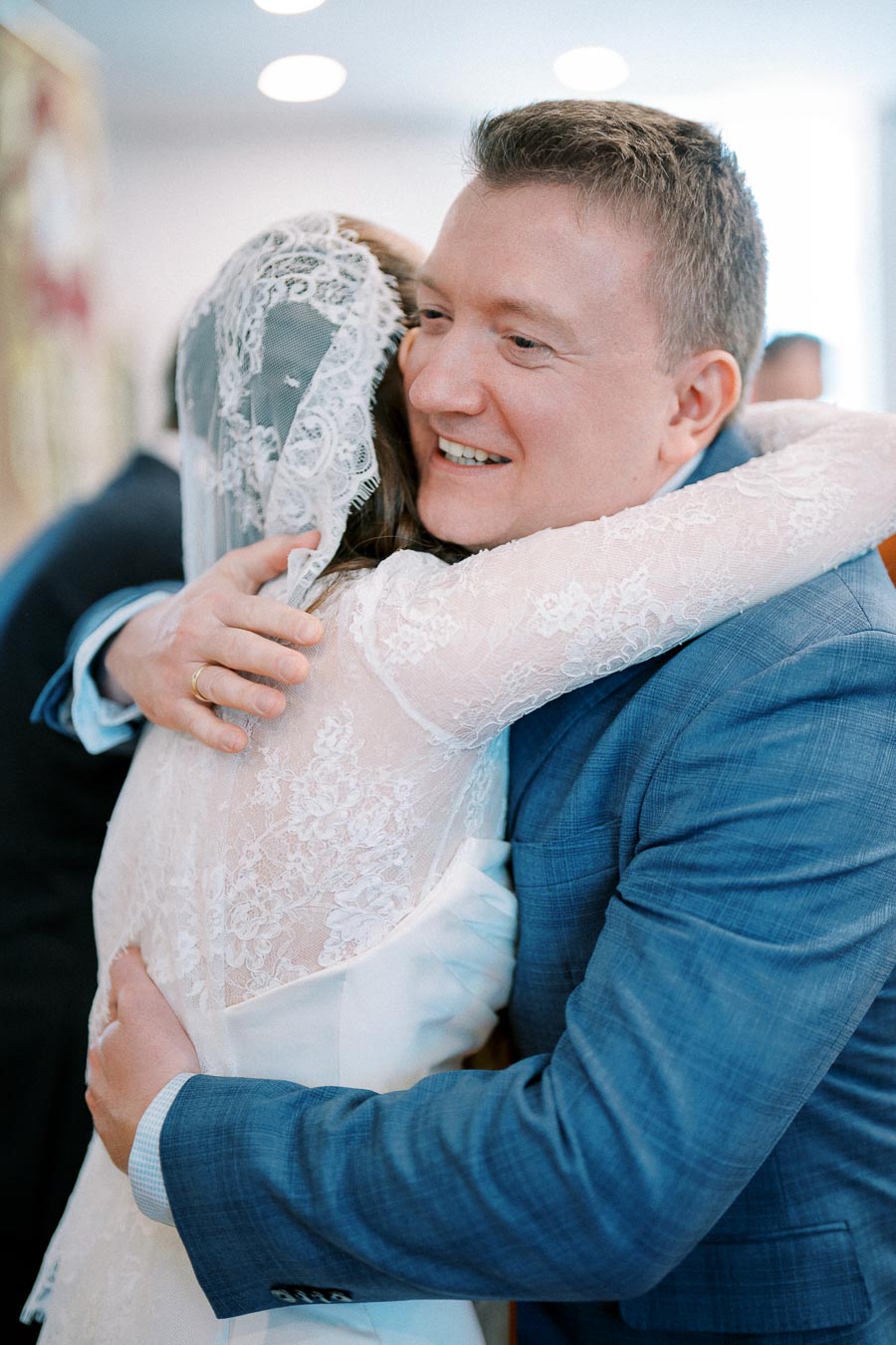 A bride and groom joyfully embrace in a wedding setting, with the groom wearing a blue suit and the bride in a lace wedding dress.