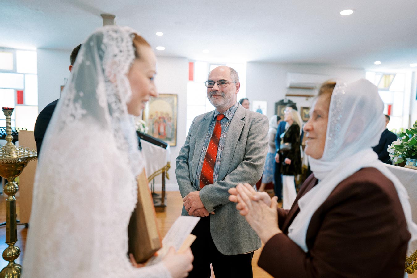 A bride in a lace veil holding a book smiles at an elderly woman with a white scarf inside a church setting, with a man in a suit observing the interaction.