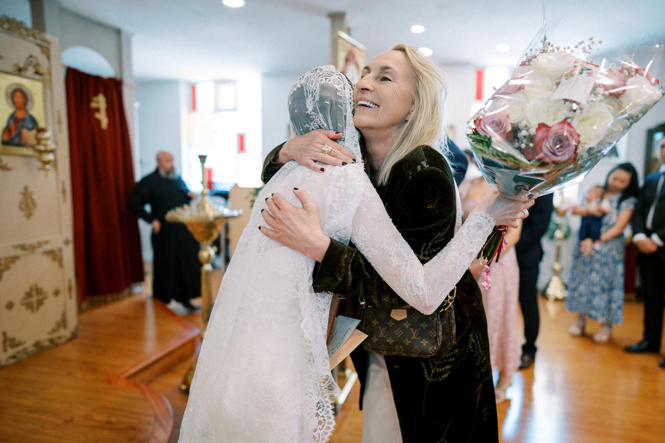 A bride in a lace dress and veil hugs a smiling woman holding a bouquet of flowers during a joyful celebration in a church setting, with onlookers in the background.