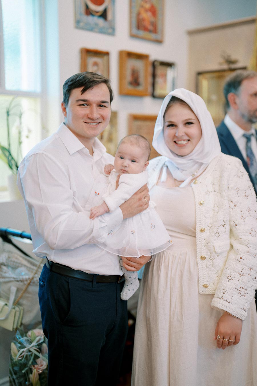 A happy family posing for a photo with their baby dressed in white at a special occasion indoors, decorated with religious artwork in the background.