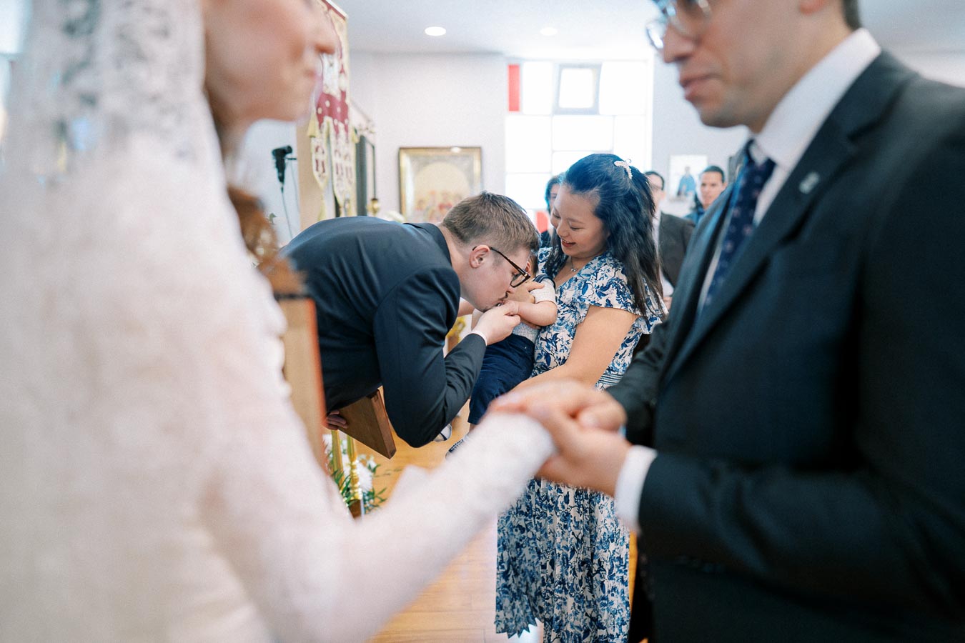 A man in a suit kisses a baby's hand held by a woman in a floral dress during a ceremony, while a couple in wedding attire holds hands in the foreground.