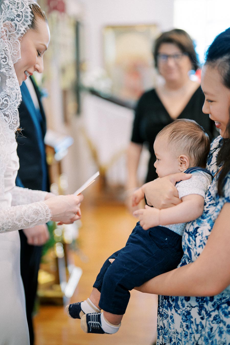Bride in lace dress smiling at a baby held by a woman, during a wedding ceremony.